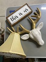 Photo showing three home decor items: beige lamp shade with fringe, white deer head with gold antlers, and wooden wall plaque reading 'this is us'