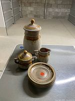 Lot overview photo showing all six pottery pieces arranged on a metal table; includes tall lidded vase, three plates stacked, sugar bowl with lid, and milk jug