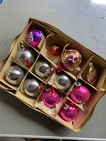 Top view of 12 antique Shiny Brite Christmas ornament bulbs in pink, silver, and gold, arranged in a cardboard box with compartments.