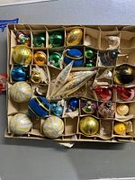 Top view of the original packaging box containing multiple assorted antique Christmas bulbs separated by cardboard compartments, showing a variety of colors and shapes including green, gold, blue, and red bulbs, as well as a large silver elongated ornament.