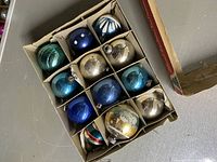 Top-down view of the set of 12 ceramic Christmas bulb ornaments neatly placed in original divided cardboard box. Colors include blue, silver, turquoise, and greenish hues with painted decorative designs.