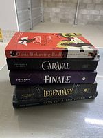 Stack of five fantasy books: Gods Behaving Badly, Caraval, Finale, Legendary, Son of a Trickster on a gray surface in a storage setting.