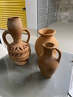 Three outdoor clay pottery items on a table: tall amphora with cutout leaf pattern, rounded pot with holes, and narrow-neck jug.
