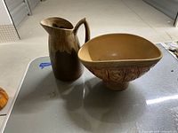 Shows a brown drip glaze ceramic pitcher next to a light brown pedestal vase with embossed patterns.