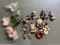 Top-down photo of all ornaments laid flat on grey background showing variety of figures including Santa Claus, snowmen, angels, green stocking, clear dove, nutcracker, and wreath.