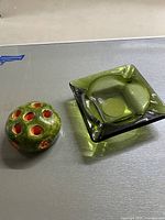 Photo showing two vintage green glass items: a round flower frog with orange-red interior holes, and a square ashtray with green translucent glass and a rounded handle.