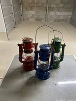 Three vintage-style oil lamps in red, blue, and green metal with glass enclosures and hanging handles, displayed together.
