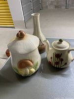 Photo showing the mushroom jar, Quebec pottery vase, and Buchanan teapot grouped on a table in a storage area.