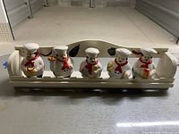 Full view of the five ceramic chef spice containers seated in the cream-colored wooden shelf with painted fruits and vegetables on the back panel.
