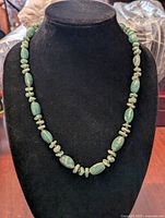 Green jade necklace displayed on a black stand, showing the alternating oval and disc-shaped beads.