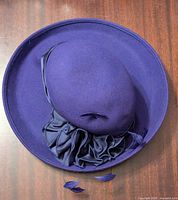 Top view of purple felt hat showing round crown, wide brim, and purple decorative gathered fabric band with feather accents