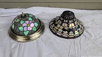 Two stained glass light fixture shades on white cloth background, one floral design in brass-colored frame, the other geometric cream and iridescent glass in dark metal frame.