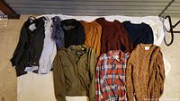 Photo shows nine men's clothing items laid out: sweaters in multiple colors, dress shirts including plaid, and jackets including an olive green bomber jacket.