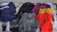 Overview photo of men's clothing lot, showing multiple tops including Adidas sweatshirt, Minnesota Wild hoodie, orange and yellow t-shirts, dark knit sweater, and bomber jacket.