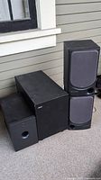 Two black Aiwa SX-N330 speakers and two subwoofers (one Jamo SUB200 and one TEAC MC-DX32i) pictured outside near a wall, showing front sides with fabric covers on speakers and port on Jamo subwoofer.