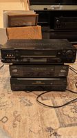 Three black VHS players stacked on carpet showing overall front and side of devices with boxes and shelves in background.
