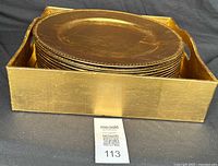 Stack of 14 round gold decorative trays with beaded edges inside a rectangular gold tray holder, angled top view.