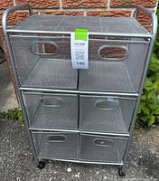 Photo of gray metal framed rolling storage cart with six mesh drawers, placed outdoors against a brick wall.