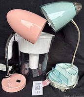 Image shows Brita water filter pitcher with white lid and filter centered between two table lamps, one pastel pink with flexible arm and round base, the other light teal with flexible arm and base containing translucent drawers.