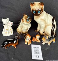 Lot of ceramic animal figurines displayed on black fabric background, includes two large Siamese cat figurines, one small white cat group, one black and brown dachshund, and four small dog figurines.