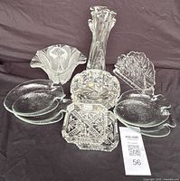 Clear glass pieces grouped on fabric including one tall vase, two square candy dishes, a napkin holder, and three uniquely shaped dishes (shell, leaf, flower).
