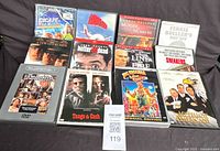 Photo showing 12 DVD movies spread out on a dark surface including titles like Escape with Honor, Alligator, Murder in Numbers, Ferris Bueller's Day Off, Tango & Cash, and others.