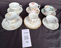 Six porcelain teacups and saucers displayed on black surface, showing various floral designs and the lot tag.