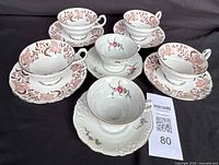 Six teacups and saucers displayed on dark background: four with brown and white floral leaf pattern and two with pink rose floral patterns including gold trim