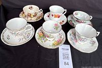 Full view of six porcelain teacups and saucers arranged on black fabric, showcasing various floral and fruit designs.