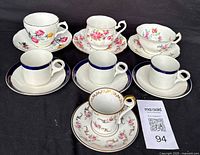 Six porcelain teacup and saucer sets displayed on a black cloth. Three sets have floral designs with pink flowers and gold trim, and three sets have cobalt blue borders on white porcelain.