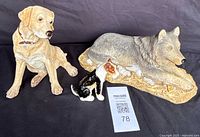 Three animal figurines: large wolf figurine lying on base, yellow Labrador dog statue, and small multicolored dog figurine on black background.