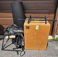 Photo showing a set of exercise equipment including a rolled black mat, metal chin-up bar placed on a wooden plyo box labeled 30, a black metal plyo box, and a black resistance parachute on a stand.