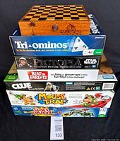 Photo showing stack of 7 board games with a wooden chess/checker box on top: Tri-Ominos, Pictopia Star Wars, Beat the Parents, Clue, Mouse Trap, and Spin Master Game.