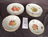 Grouped view of the fruit motif tableware set including dinner plates, salad plates, and small bowls with gold trim on a dark cloth background.