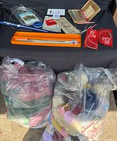 Two large bags filled with assorted yarn skeins of various colors including pink, cream, green, and more, alongside knitting needles and related tools arranged on a black cloth.