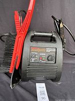 Front view of the black MotoMaster Eliminator battery charger with red and black clamps, attached black brush visible on the side.