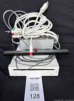 White Nintendo Wii console with front disc drive, black sensor bar placed on top, power adapter and AV cables coiled and arranged behind the console.