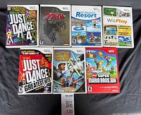 Photo of seven Nintendo Wii game cases arranged on a dark fabric background: Just Dance 4, The Legend of Zelda: Twilight Princess, Wii Sports Resort, Wii Play, Just Dance Greatest Hits, Star Wars The Clone Wars: Lightsaber Duels, New Super Mario Bros. Wii.