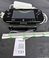 Photo showing the black Nintendo Wii U GamePad resting on top of the black Wii U console with front slot for game disc, along with a white sensor bar wrapped with green tape, power supply cable visible.