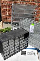 Photo showing a black plastic organizer with 24 small clear drawers and two metal mesh organizers with horizontal open shelves, placed outdoors against a brick wall.