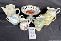 Seven ceramic tea time pieces arranged in a row, showing variety of designs, including teapot, sugar bowls, creamers, and treat plate with colorful floral decorations and pierced edges.