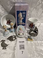 Vintage figurine lot with various items including two Age 2 figurines holding gold hearts, Precious Moments figurine boxed, baby and mouse figurines, teddy bear cup and bowl, and three pewter trinkets, displayed against a patterned fabric backdrop.