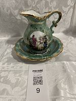 Front view of the green jug with gold trim and classical scene, showing ornate handle and pedestal base.