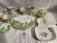 Full lot view showing German Lustre creamer and sugar bowl in yellow lustre glaze; green bear mini tea set with visible wear; ceramic serving dishes with painted designs of countryside house, trees, and bears; olive and square serving dishes with painted fruit and bird motifs.