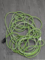 Photo showing a green 3-prong outdoor extension cord coiled and slightly tangled on a dark wooden floor.