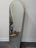 Full view of tall wall mirror standing against a wall showing its arched top and full length reflective surface, gold-toned slim frame visible.