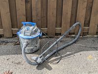Image of the Craftsman shop vacuum showing front angle with hose and nozzle attachment, placed on pavement in front of wooden fence.