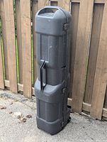 Black hard case golf club travel caddy standing upright against a wooden fence, showing the handle and body shape.