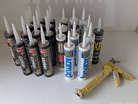 Front view of multiple sealed caulking tubes in black, white, and blue labels alongside a yellow used caulking tool.
