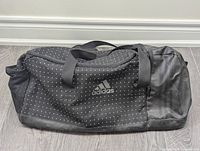 Front and top view of a black Adidas gym/duffel bag with white dots and leather bottom panel, showing handles and Adidas logo on front pocket.